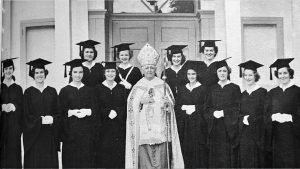 A black and white photo shows a Catholic bishop dressed in white, with the staff, surrounded by female graduates in cap and gown.