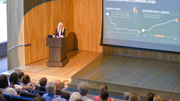 Tama Duffy Day stands on stage at a podium with a slideshow behind her during the Keegan Lecture.