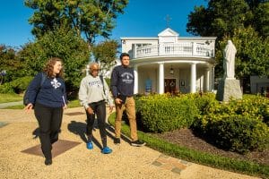 Two women and a man with sweatshirts walk along a path in front of a chapel.