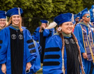 Two female college graduates walk, smiling, clothed in cap and gown. 
