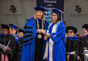 President Irma Becerra, in doctoral cap and gown, poses shaking hands with a female graduate on the graduation stage. 