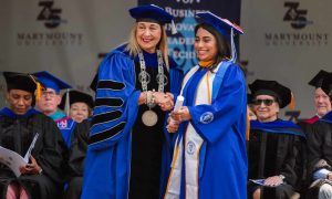 Woman in cap and gown receives diploma from a woman in doctoral hood and gown.