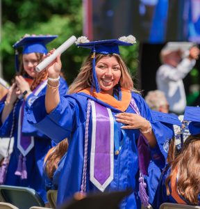 A woman with long hair in graduation cap and gown smiles and waves her diploma at the Marymount graduation ceremony.