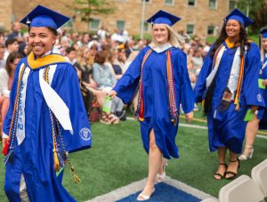 Three graduates in cap and gown walk across a field, preparing to cross the graduation stage.
