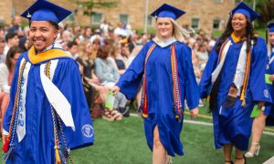 Three graduates in cap and gown walk across a field while an audience sits behind them.