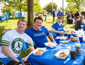 A group of alumni dressed in Saints Lacrosse shirts sit down and eat hamburgers and grilled food at a table.