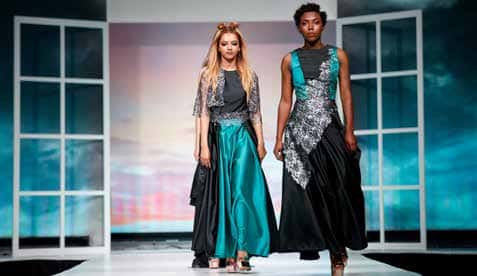 Two student models in long blue, grey, and black gowns walk down a runway