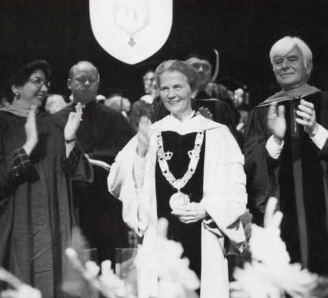 A woman in a robe and with a large medal waves while the men and women around her, also clothed in academic robes, applaud.