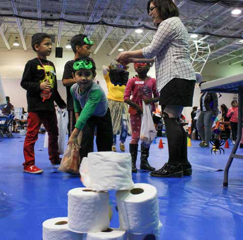 A child with a mask and costume throws a ball at a toilet paper pyramid.
