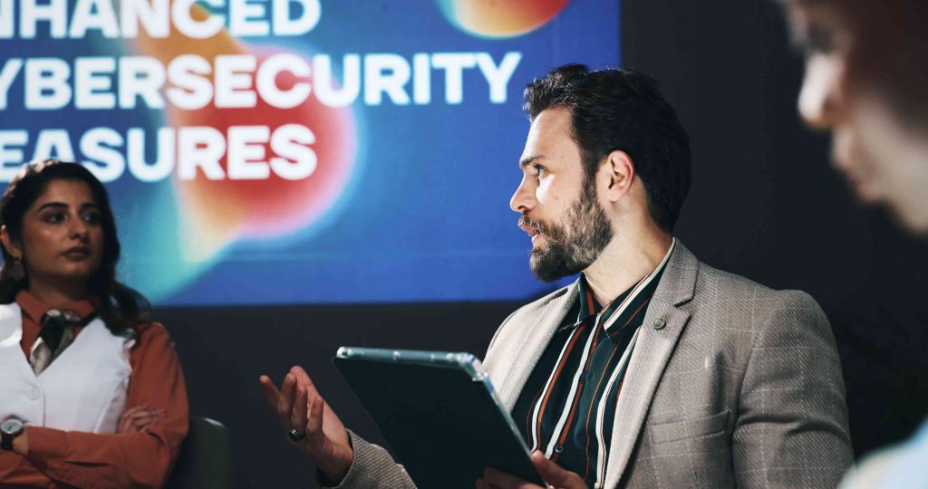 People appear to sit on a stage. One person holds a tablet. In the backgrounds the words Enhanced Cybersecurity Measures appears on a presentation screen.