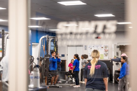 Students in the weight room. One stands under the barbell and prepares to raise it above his head.
