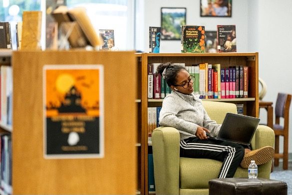 A student sits in a green chair in front of a bookshelf and works on her laptop.