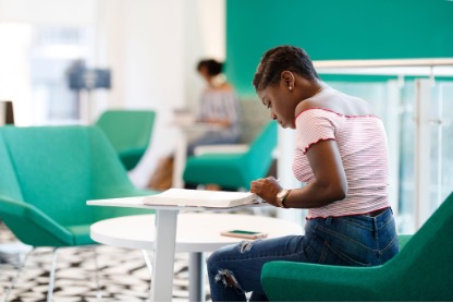 A student sits on a green lounge chair and leans across a table, reviewing her notebook