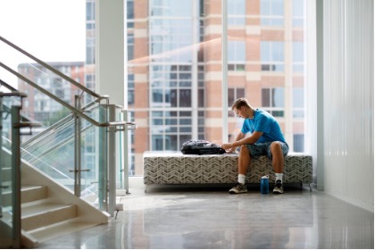 A student sits on a long lounge bench in front of a floor to ceiling window and pulls something out of his backpack