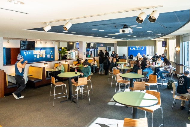 Students sit in a large restaurant-style cafeteria with booth tables. A projector is attached to the ceiling so that they can have large presentations or slideshows on the wall.