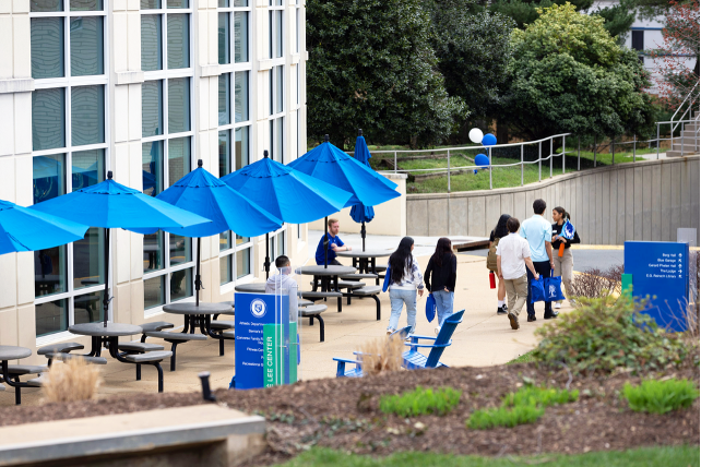 Students walk by and sit at picnic tables with blue umbrellas on a patio in between a garden and a building