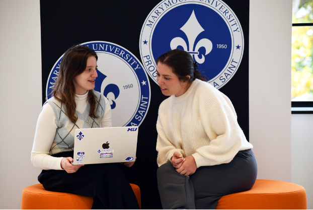 Two students sit on a bench and talk to each other while leaning over a laptop. The background has Marymount logos with fleur-de-lis