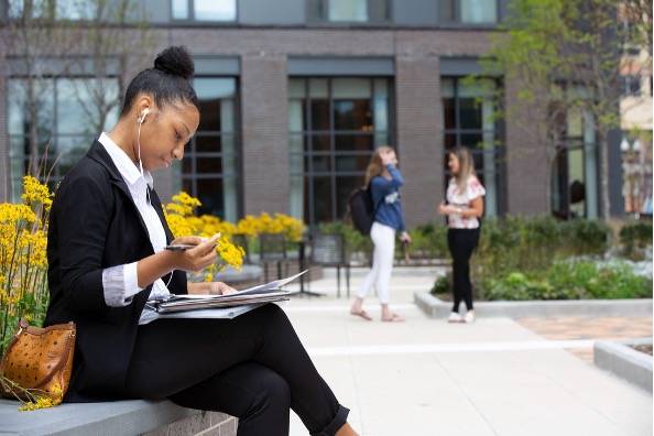 A student sits on a bench working on papers while two other students stand talk to each other in the background